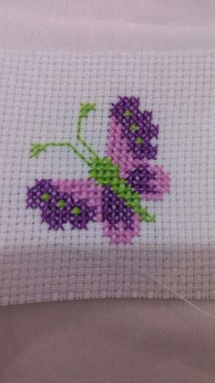 a cross stitched purple flower with green leaves on it's side, sitting on top of a white table cloth