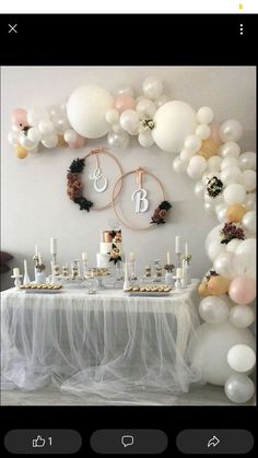 a table topped with lots of white balloons and desserts next to a wall mounted monogramme