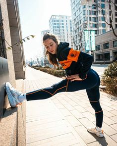 a woman stretching her legs on the side of a building while wearing running shoes and an orange vest