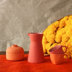 three vases and two cups sitting on an orange table cloth next to a large flower arrangement