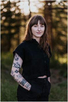 a woman with tattoos on her arm standing in front of trees and looking at the camera