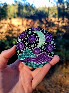 a hand holding up a purple and green beaded brooch with a crescent moon on it