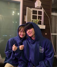 two people sitting on a bench in front of a house wearing blue hoodies and smiling at the camera