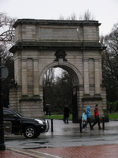 people walking in front of an arch on a rainy day with cars parked near by