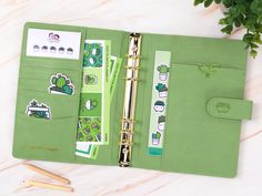 an open green planner with stickers and pens on the side next to a potted plant