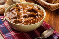 a bowl of beef stew on a table with bread in the background and a spoon next to it