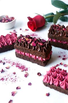 four pieces of chocolate cake with pink frosting and rose petals on the table next to it