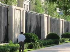 a man standing on the sidewalk in front of a row of trees and bushes, with his back turned to the camera