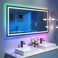 two sinks in front of a large mirror with lights on the wall above them and flowers