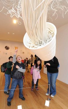 a group of people standing in front of a white vase filled with noodles on top of a wooden floor