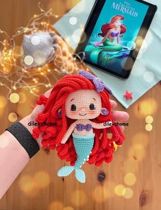 the little mermaid doll is being held up by someone's hand, next to an ipad