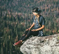 a young woman sitting on top of a rock next to a forest filled with trees