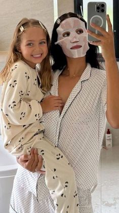 a woman in pajamas holding a child wearing a face mask and taking a selfie