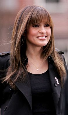 a woman with long hair wearing a black jacket and smiling at the camera while standing in front of a brick building