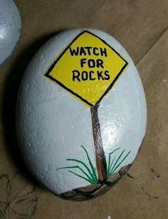 a painted rock with a yellow sign that says watch for rocks on it and grass growing underneath