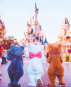 three stuffed animals are standing in front of the castle at disney world, one is wearing a pink bow tie