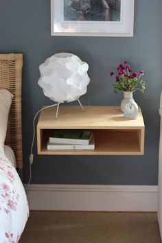 a small table with a lamp on it next to a bed and flowers in a vase