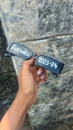 a person holding up two black glasses with the word farewell written on them in front of a stone wall