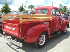 an old red truck parked in a parking lot next to other cars and people looking at it