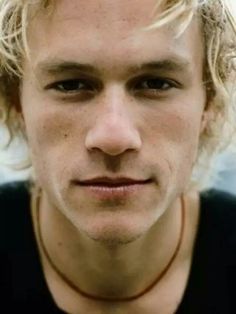 a young man with blonde hair wearing a black shirt and looking at the camera while standing in front of a cityscape