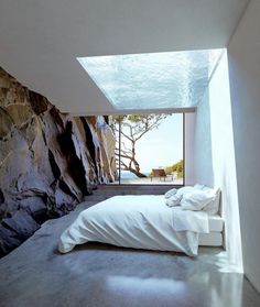 a bedroom with a large window overlooking the water and rocks on the wall, along with a bed that is covered in white linens