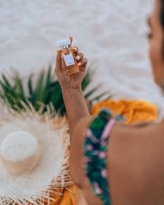 a woman holding up a bottle of perfume next to a palm leaf and white hat