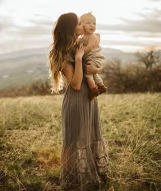 a woman holding a baby in her arms while standing on top of a grass covered field