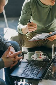 two men sitting at a table with notebooks and coffee in front of them by an open laptop computer