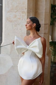 a woman in a short white dress with a large bow on her head standing next to a wall