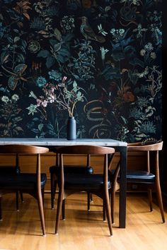 a dining room table with chairs and a vase on top of it, in front of a floral wallpaper