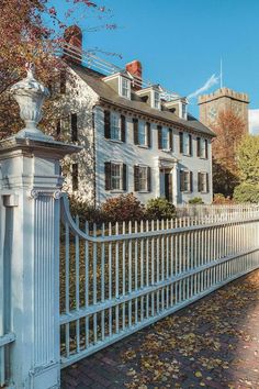 a white picket fence in front of a large house with trees and leaves on the ground