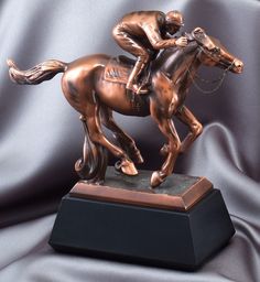 a bronze statue of a jockey riding a horse on top of a black base with a silver cloth behind it