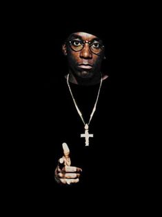 a man with glasses and a cross on his necklace pointing at the camera while standing in front of a black background