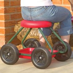 a woman sitting on top of a red and green toy car next to a brick wall