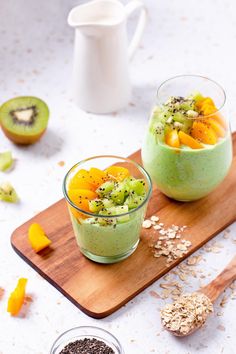 two glasses filled with green smoothie on top of a cutting board next to sliced kiwis