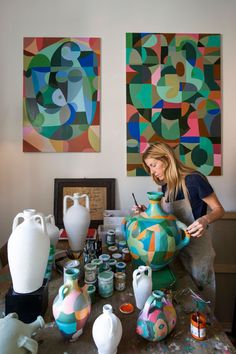 a woman is painting vases on a table with other art work in the background