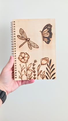 a hand holding a notebook with drawings of flowers and dragonflies on the front cover