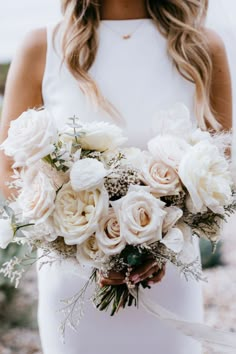 a bride holding a bouquet of white roses and greenery in her hands on the beach