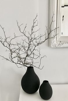 two black vases with branches in them sitting on a white table next to a mirror