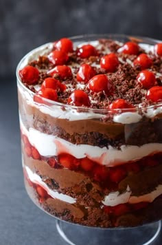 a layered chocolate cake with cherries and whipped cream in a glass dish on a black table