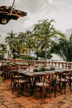 an outdoor dining area with wooden chairs and tables set up on brick flooring next to palm trees