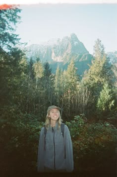 a woman standing in front of a mountain with trees and bushes on the ground, smiling at the camera