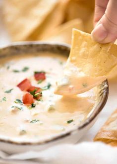 a person dipping tortilla chips into a bowl of dip with tomatoes and cheese