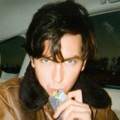 a man in a leather jacket eating something with his hand and looking at the camera