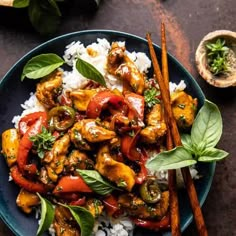 a blue plate topped with chicken and vegetables on top of rice next to wooden chopsticks