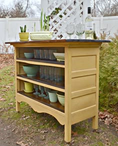 an outdoor kitchen island with pots and bowls on it's shelf, in front of a white picket fence