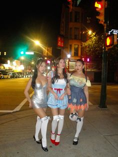 three women dressed up in costumes standing on the side walk at night with traffic lights behind them