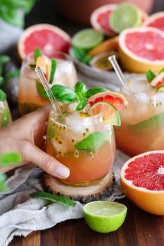 a person holding up a grapefruit cocktail in front of other glasses and fruit