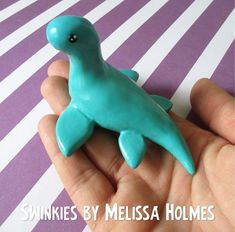 a hand holding a small blue plastic toy dinosaur in it's palm, with the words swinkies by mellisa holmes above it