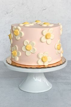 a pink cake with white and yellow flowers on it, sitting on a pedestal in front of a wall